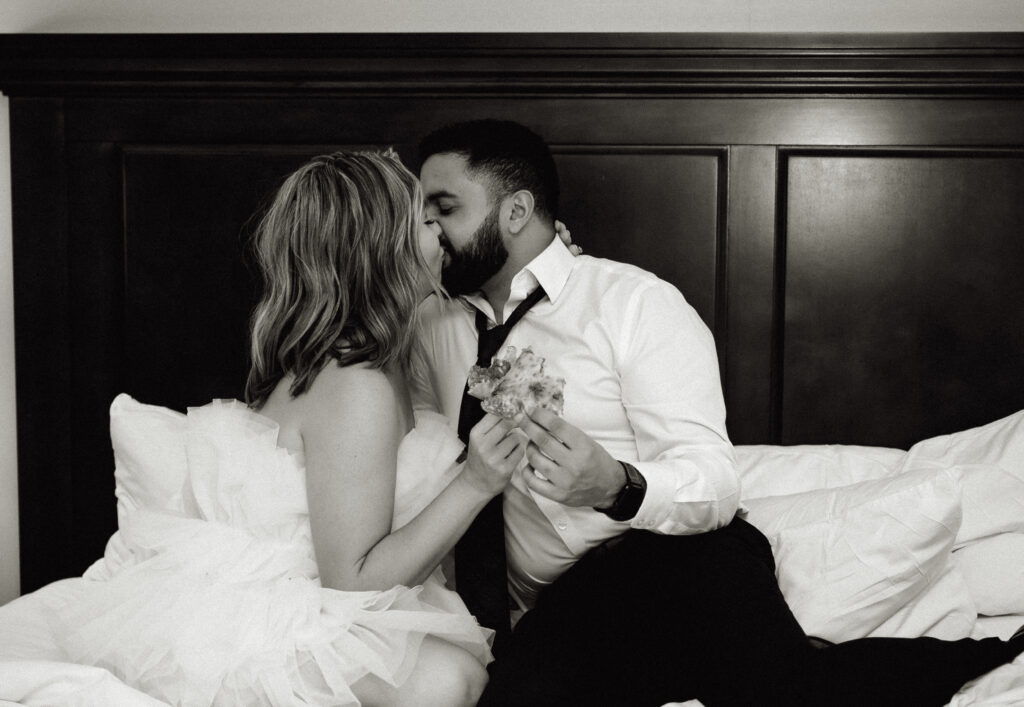 Black and White photo of bride and groom kissing while sitting in bed holding slices of pizza. 