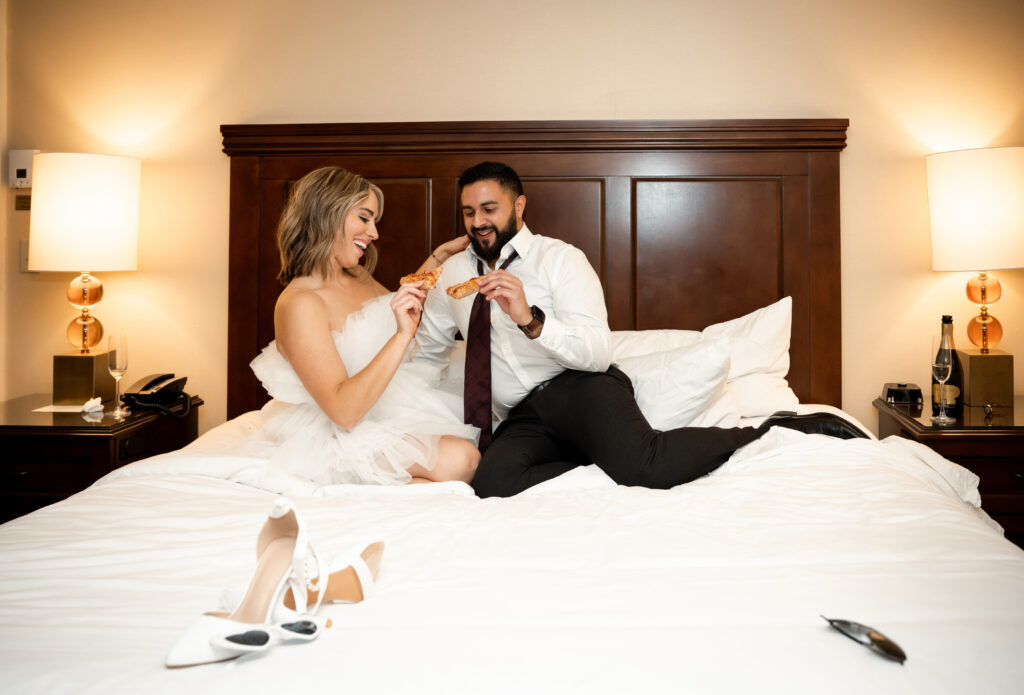 Bride and Groom sitting in bed laughing while holing slices of Pizza.