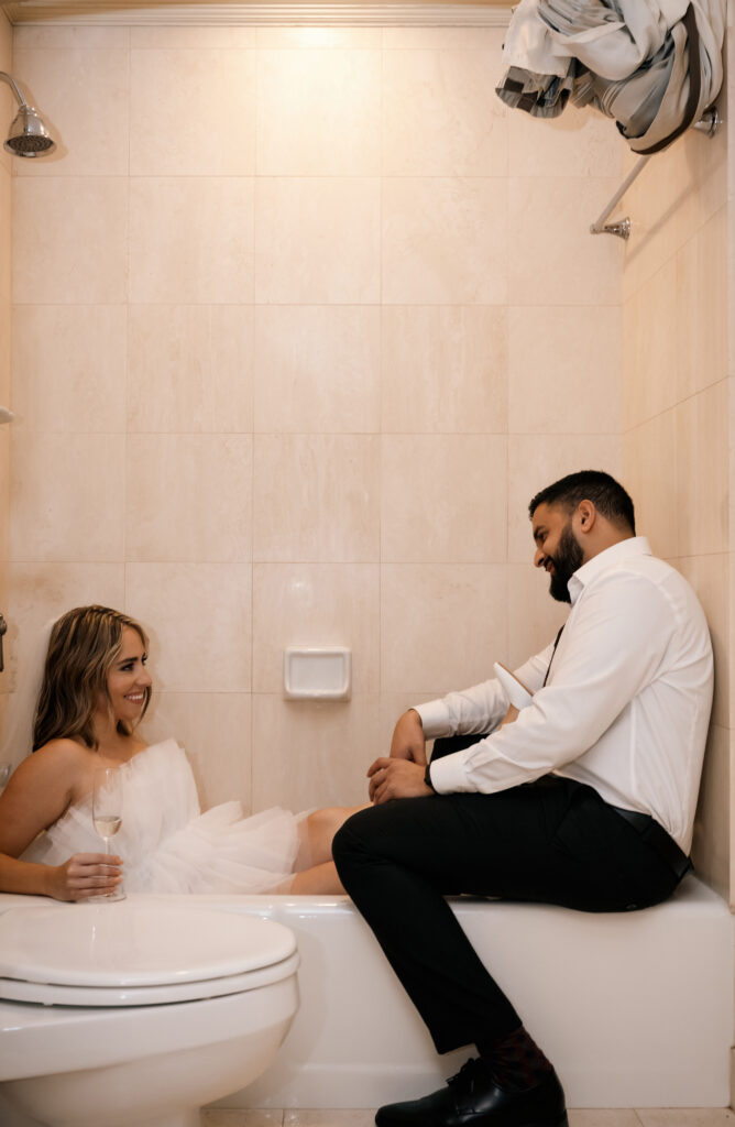 bride and groom sitting in a bathtub looking at each other while they hold champagne glasses