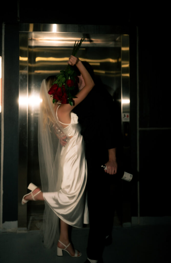 Bride and Groom kiss in front of elevator while hiding behind red roses