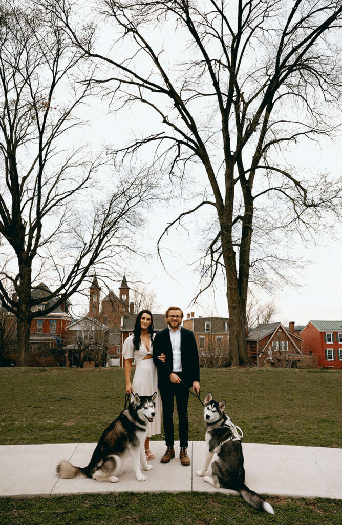 Warm documentary-style photo of couple looking at each other while holding their dogs’ leashes in an urban Pittsburgh setting.