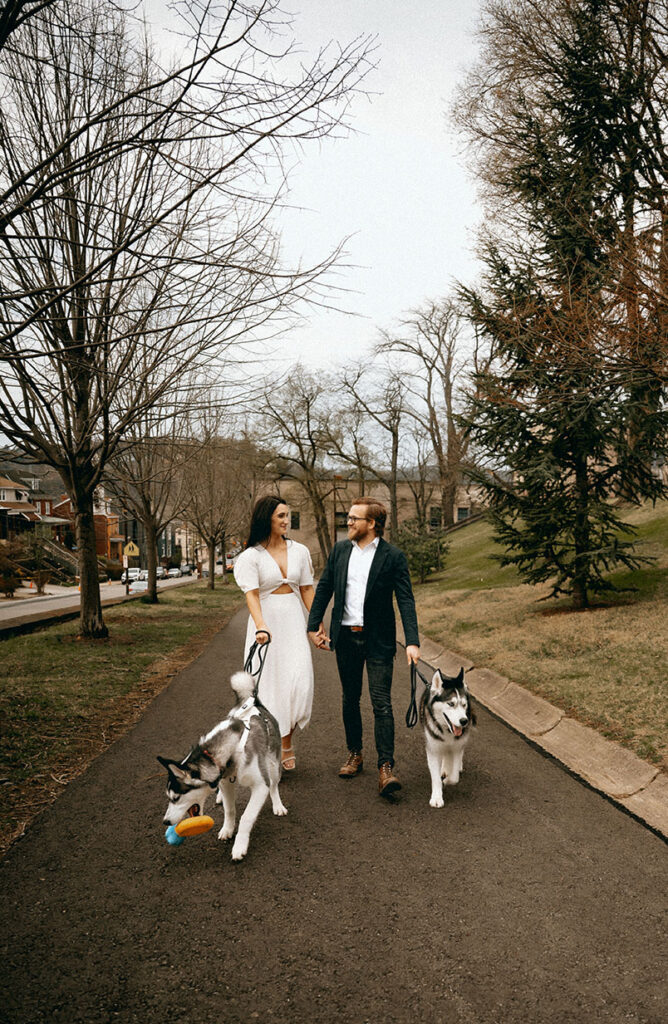 Warm documentary-style photo of couple looking at each other while holding their dogs’ leashes in an urban Pittsburgh setting.