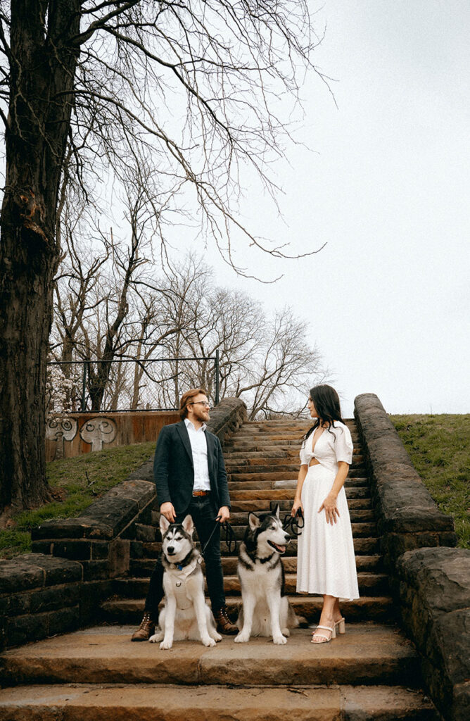 Joyful candid of couple looking at each other while their huskies play on the sidewalk, capturing a personal moment.