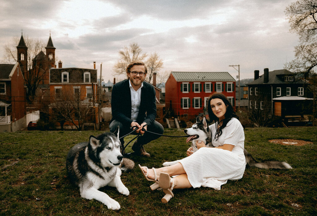 Joyful candid of couple looking at each other while their huskies play on the sidewalk, capturing a personal moment.