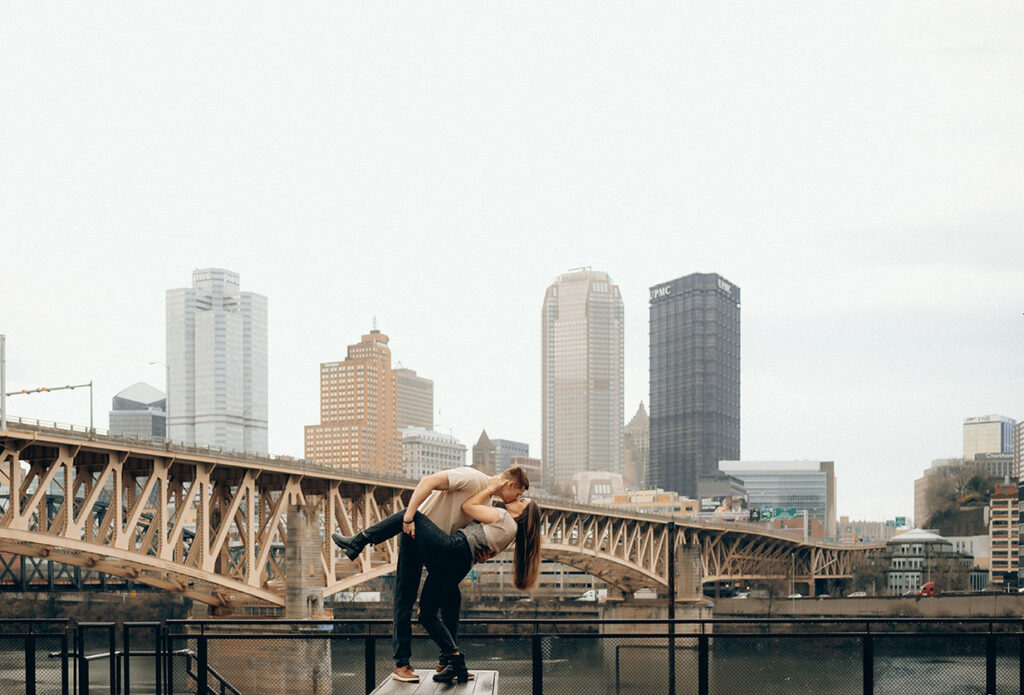 Couple kissing in front of Pittsburgh skyline during engagement session at The Highline, captured by Pittsburgh engagement photographer Natalia Baqueiro.