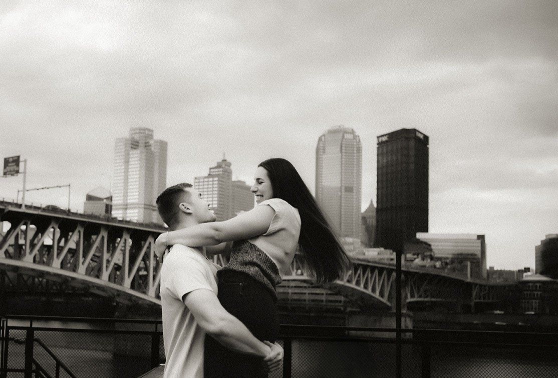 Groom-to-be lifts fiancée during their playful spring engagement session at The Highline with the city in view.