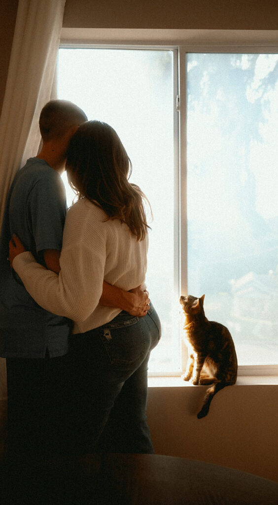 Couple Looking out the window with their cat looking at them during at home engagement