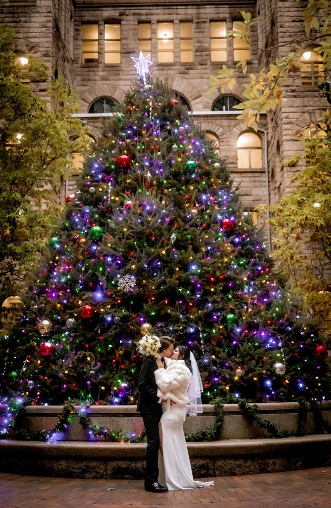 Bride wearing a white elegant winter coat next to the large natural Christmas tree in the courthouse patio.