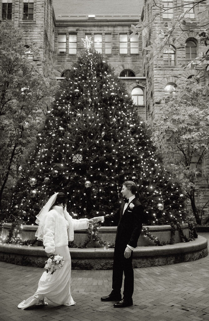 Bride wearing a white elegant winter coat next to the large natural Christmas tree in the courthouse patio.