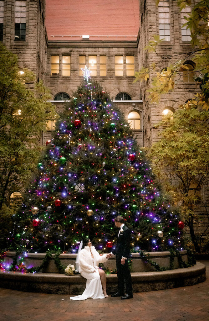Bride wearing a white elegant winter coat next to the large natural Christmas tree in the courthouse patio.