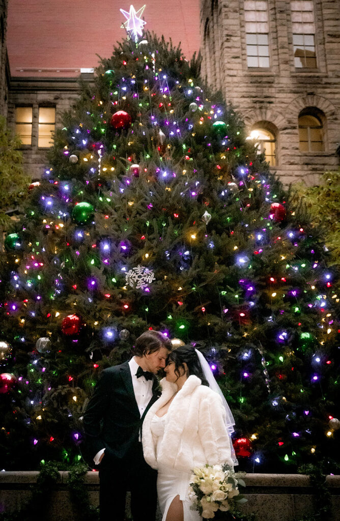 Bride wearing a white elegant winter coat next to the large natural Christmas tree in the courthouse patio.