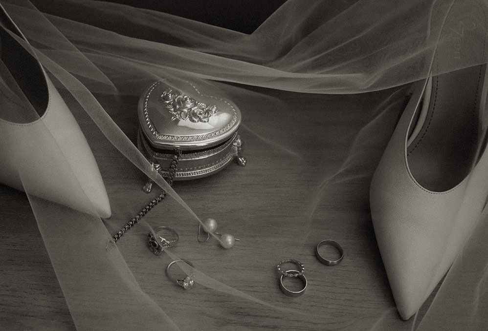Black-and-white wedding details with bridal shoes, veil, rings, pearls, and a heart-shaped jewelry box.