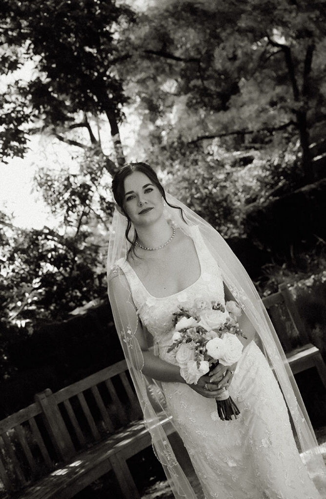 Black-and-white bridal portrait with a bouquet and flowing veil beneath the garden trees.