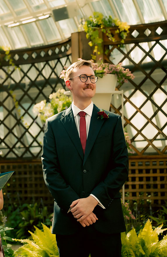 Groom in a dark green suit and red tie waiting among the gardens before the ceremony.
