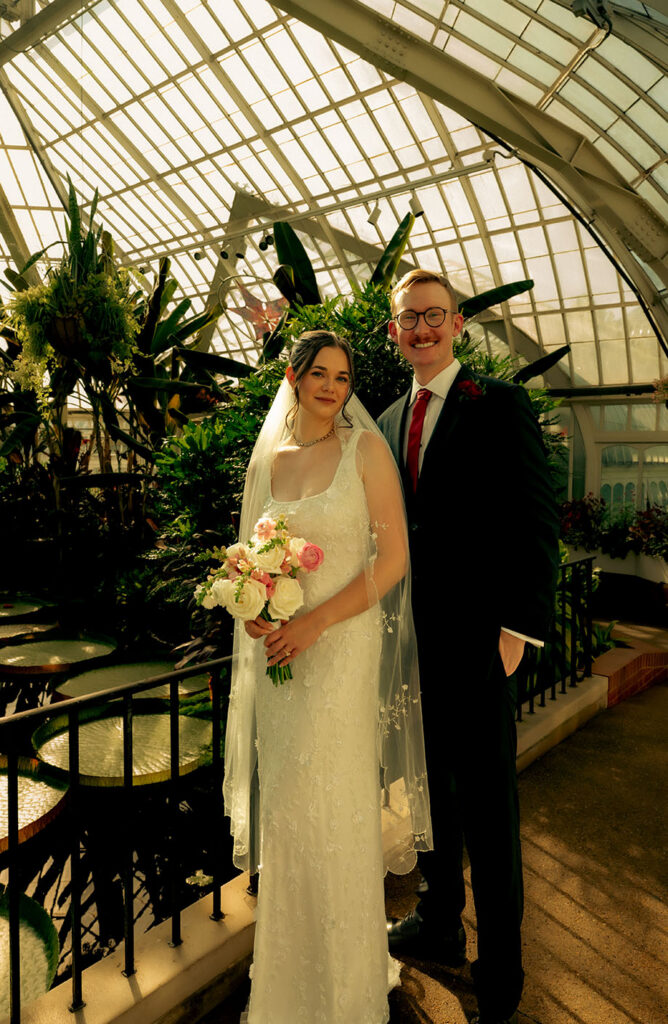 Newlyweds sharing a quiet kiss surrounded by palms and lush greenery inside the conservatory.