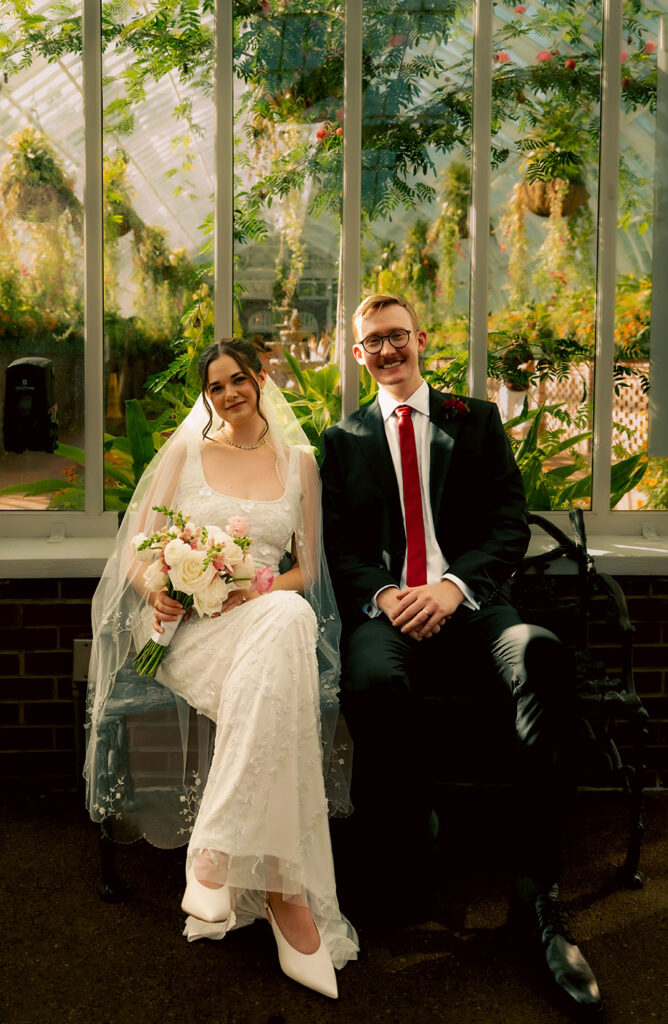 Bride and groom laughing together on a bench inside the sunlit Phipps Conservatory gardens.