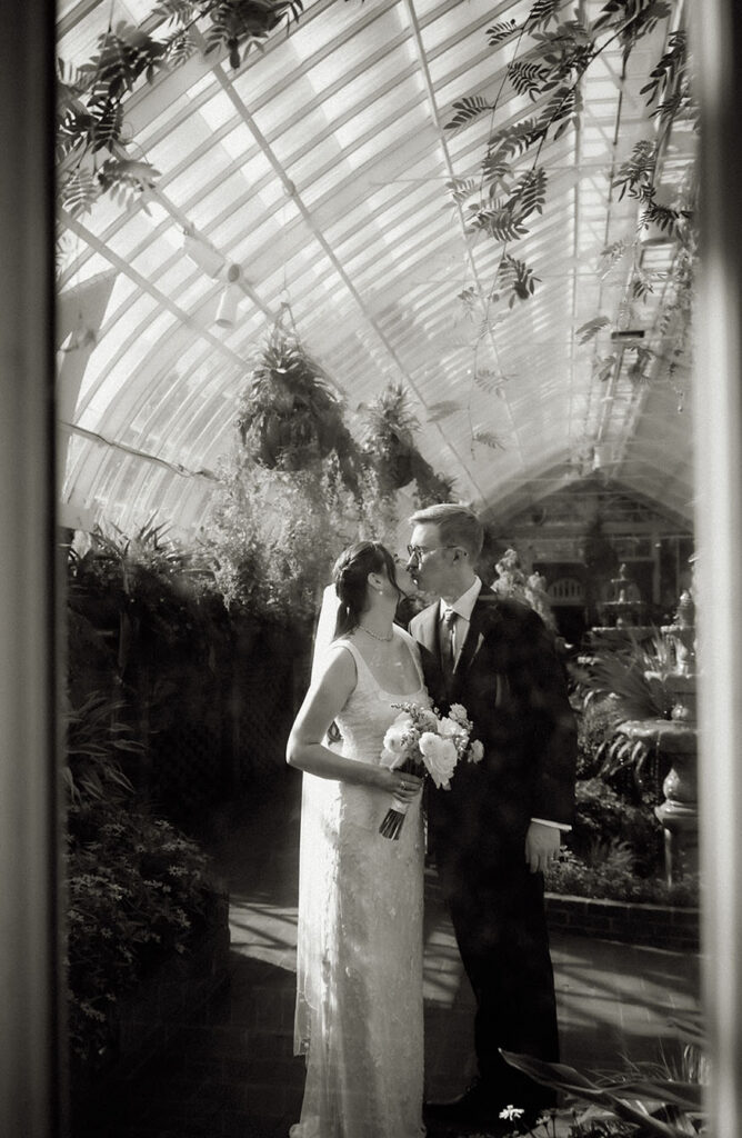 Black-and-white portrait of the newlyweds kissing beneath the bride’s veil in the glasshouse.