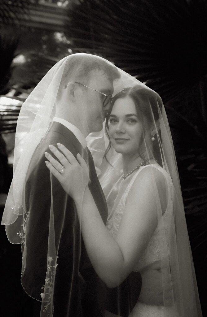 Black-and-white portrait of the newlyweds kissing beneath the bride’s veil in the glasshouse.