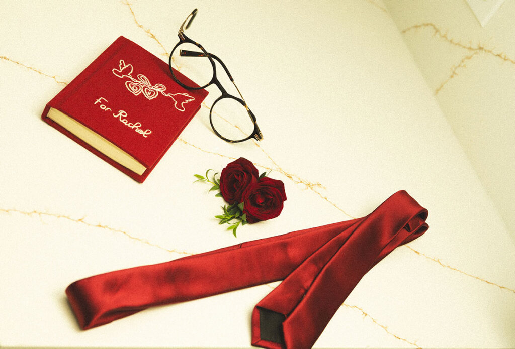 Ivory bridal shoes and jewelry arranged beside the groom getting ready with his red vow book.
