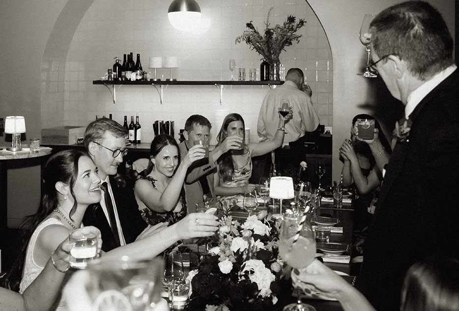 Newlyweds and guests raising their glasses together in a joyful black-and-white reception moment.